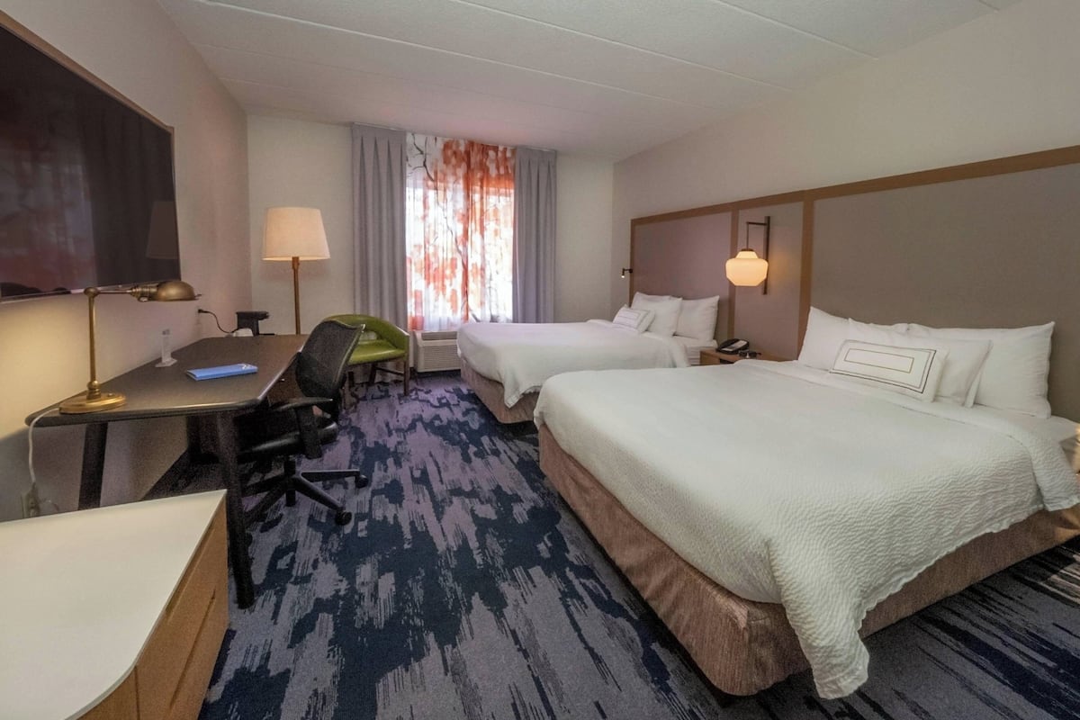 A hotel room with two beds, a desk with a chair, a TV, and a window with curtains.