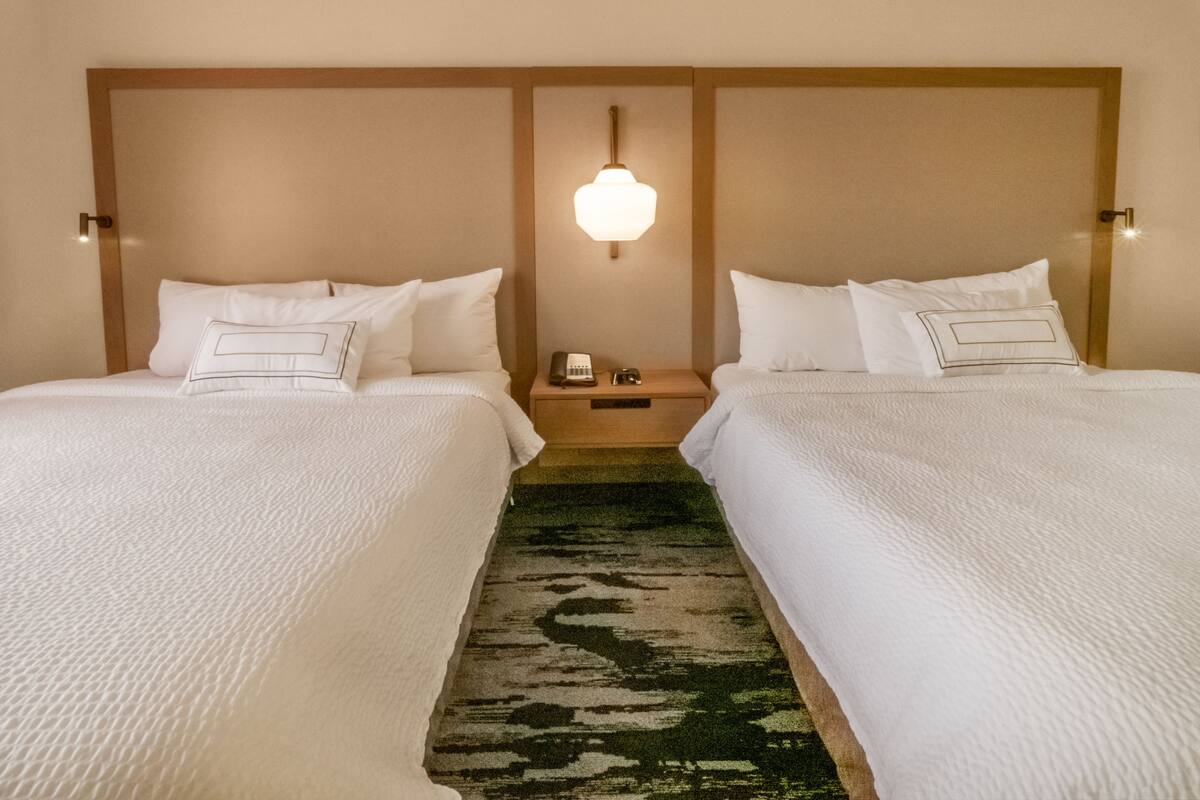 A hotel room with two beds, a nightstand with a phone, and a wall-mounted lamp.