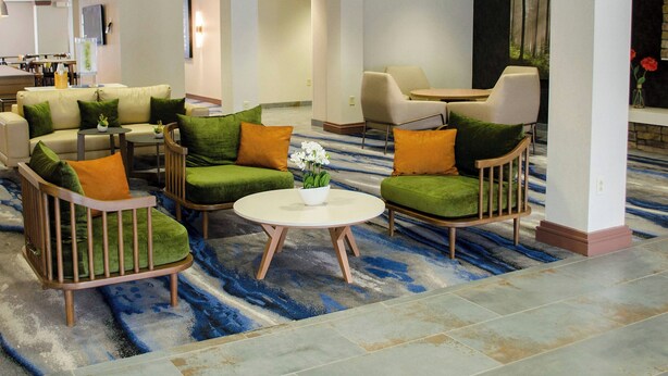 Un vestíbulo de hotel con asientos verdes y beige, una mesa redonda con un florero y un ventanal con vistas a árboles.