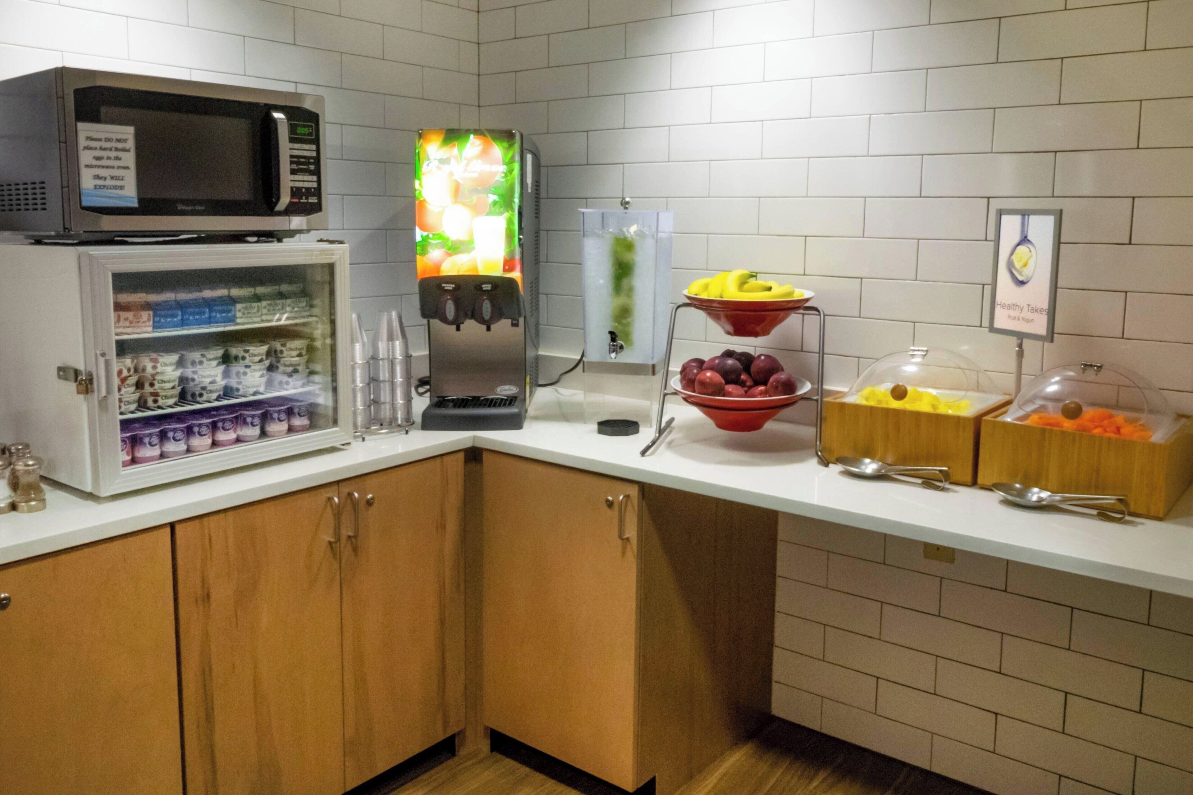Una cocina con microondas, dispensador de fruta, refrigerador y varios alimentos sobre la encimera.