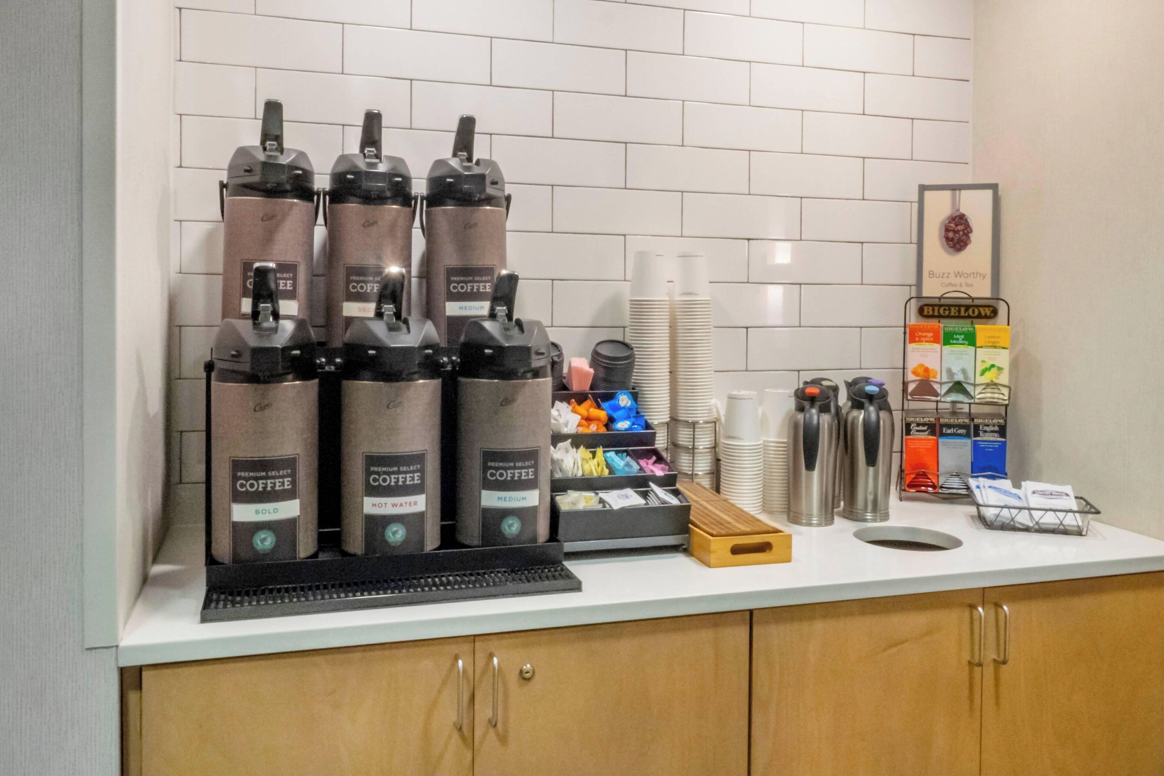 Una estación de café con varios dispensadores, una pila de vasos desechables y un menú de café en la pared.