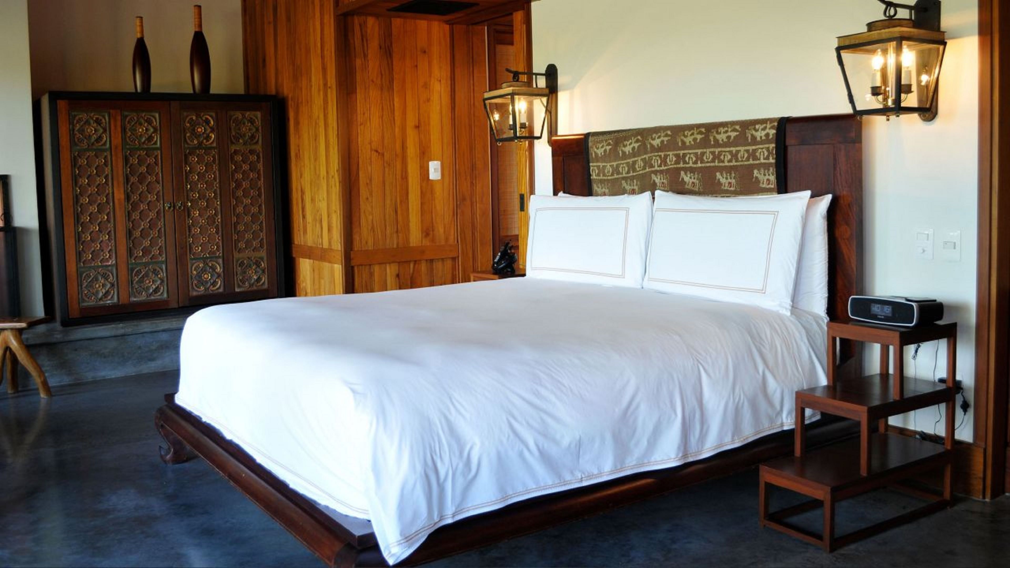 Una cama grande con ropa blanca, cabecera de madera y dos lámparas fijadas a la pared.