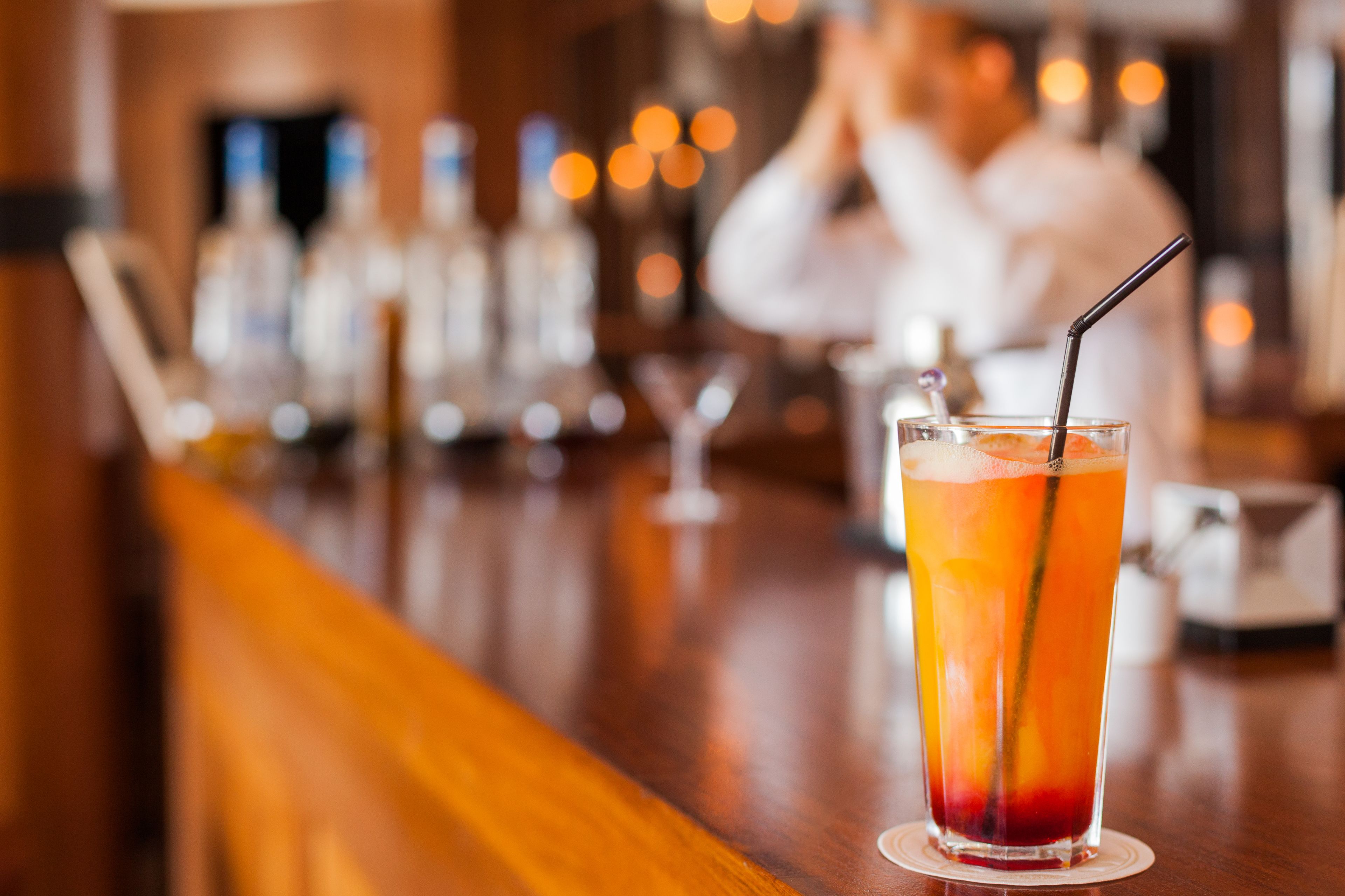 A bar with a tall glass of a layered drink, a bartender in the background, and bottles of liquor on the shelves.