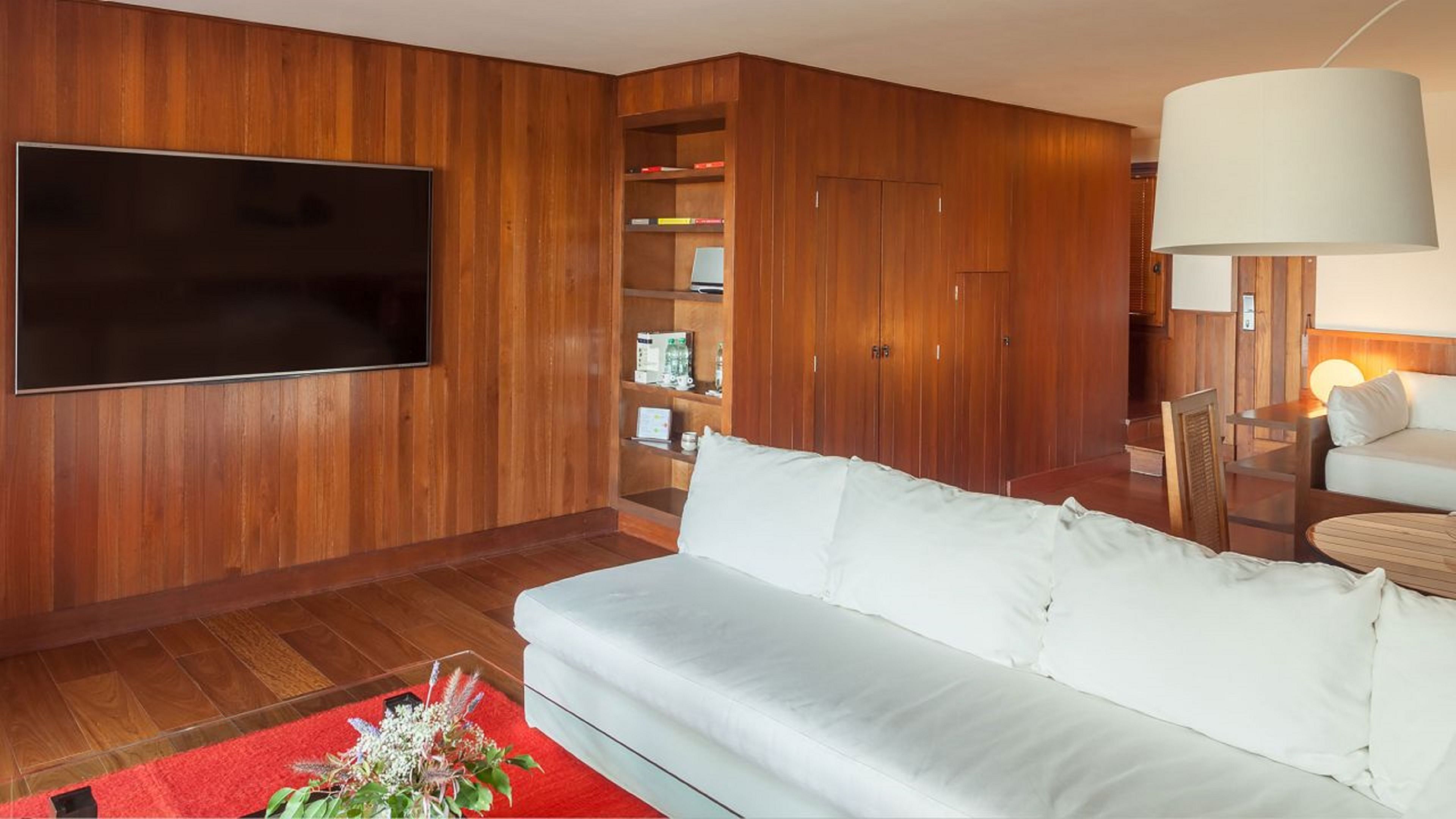 A modern living room with a flat-screen TV, a white sofa, wooden panel walls, a red rug, and a large lamp.
