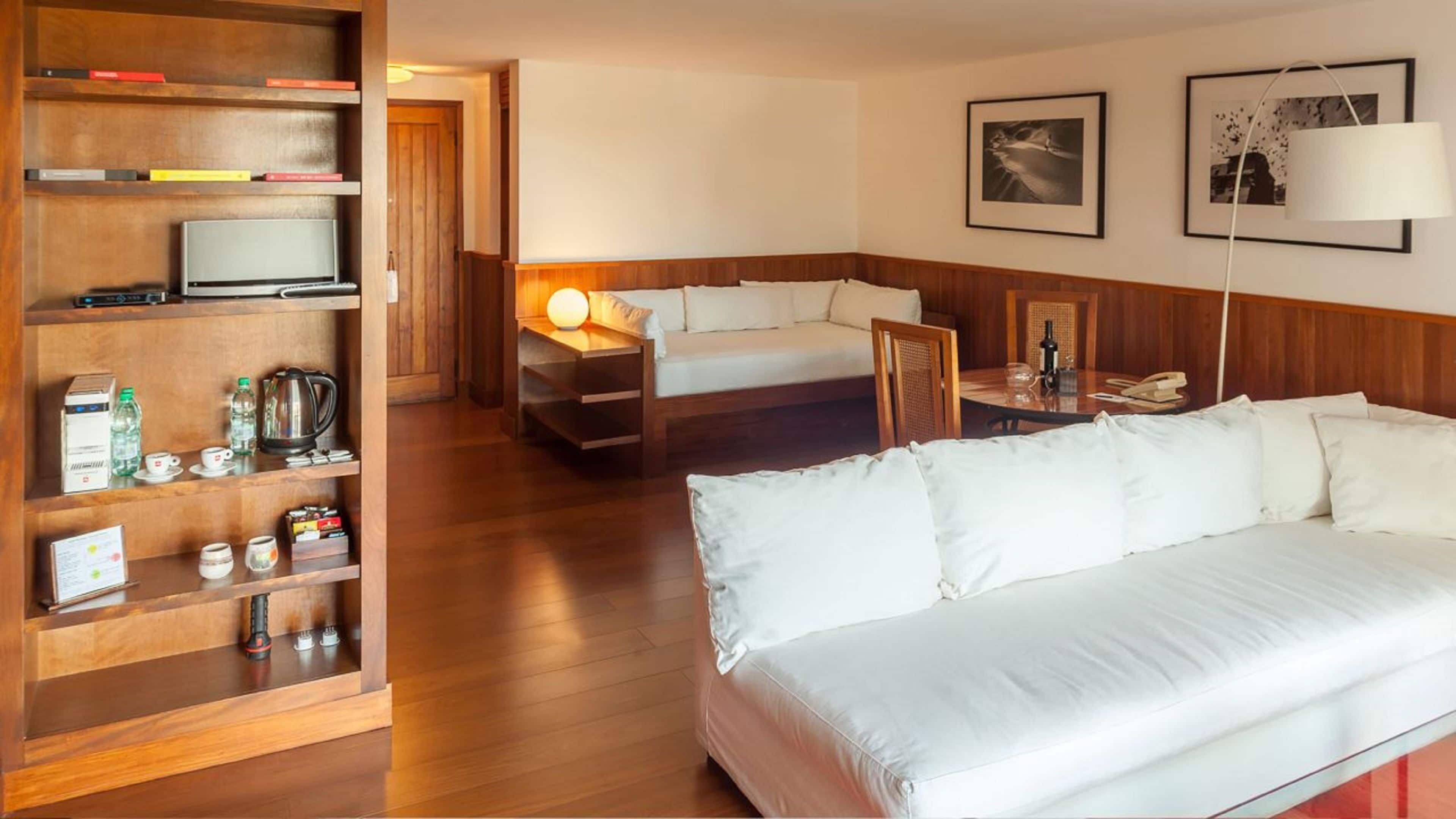 A hotel room with a large bed, a smaller bed, a wooden bookshelf, a desk with a lamp, and two framed pictures on the wall.