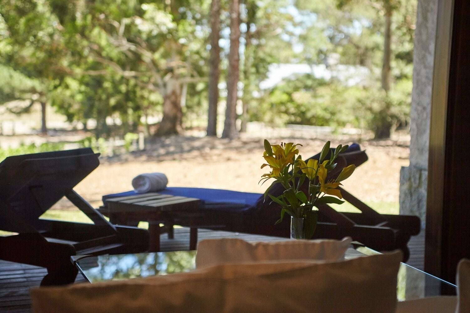 Une chaise longue et une petite table sur laquelle est posé un vase rempli de fleurs jaunes, le tout se dressant sur un fond de verdure composé d’arbres et d’une terrasse en bois.
