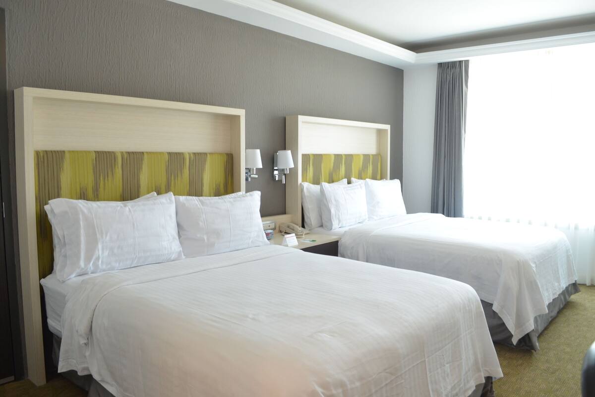 A hotel room with two beds, a bedside table with a lamp, and a window with curtains.
