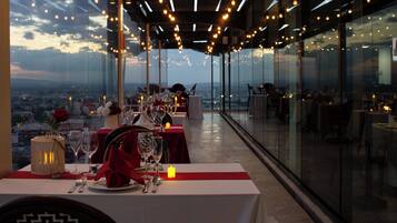 A restaurant with tables set for dining, a view of the cityscape, and a glass-enclosed ceiling with hanging lights.
