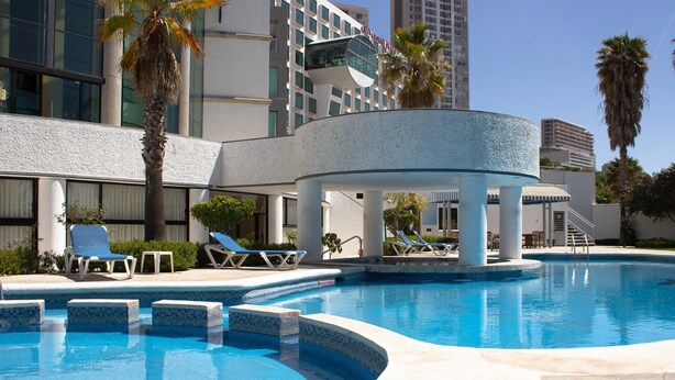 A swimming pool with lounge chairs and palm trees, surrounded by modern buildings. - Holiday Inn Queretaro Zona Diamante by IHG (Querétaro)