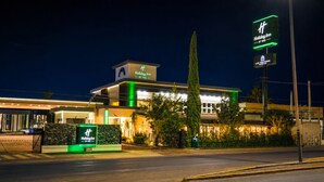 A Holiday Inn hotel with a prominent sign, a well-lit facade, and a tall cypress tree in front.