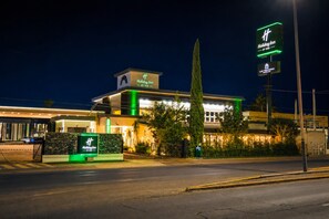 A Holiday Inn hotel with a prominent sign, a well-lit facade, and a tall cypress tree in front.