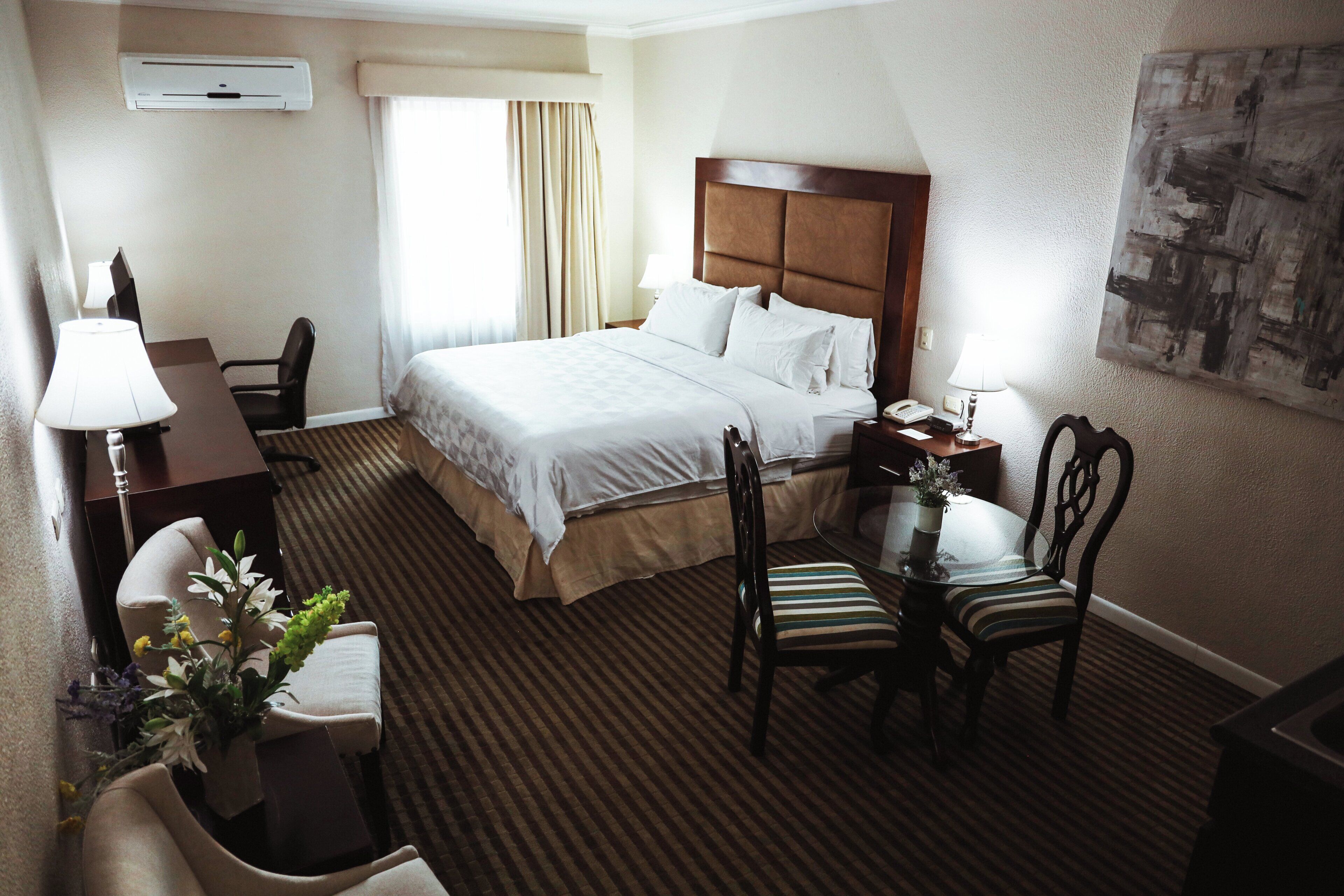 Habitación de hotel con cama, mesita de noche, lámpara, silla, escritorio y una pequeña mesa con un jarrón de flores.