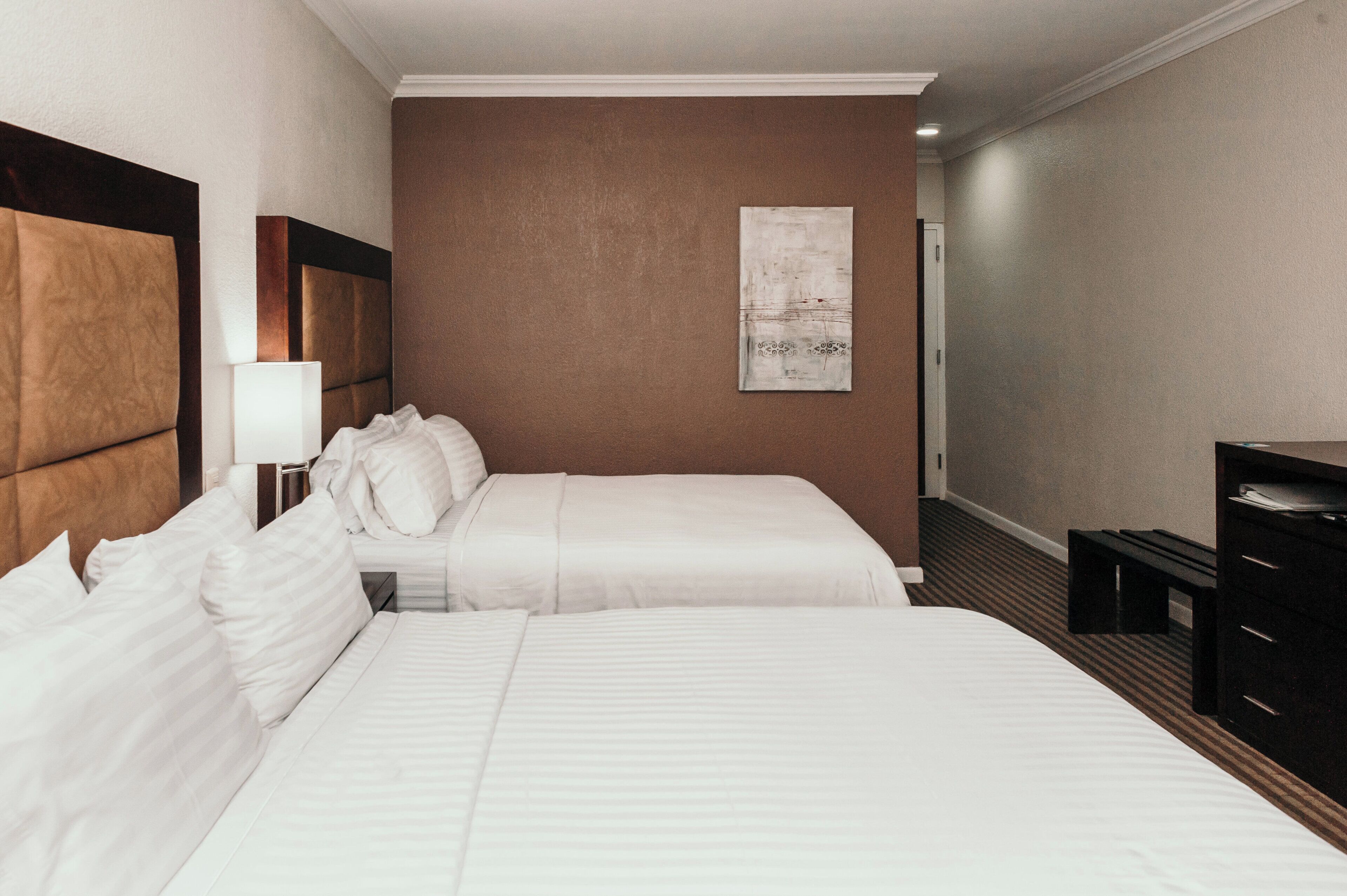 Una habitación de hotel con dos camas, cabecera, mesita de noche con lámpara, una obra de arte en la pared y un aparador.
