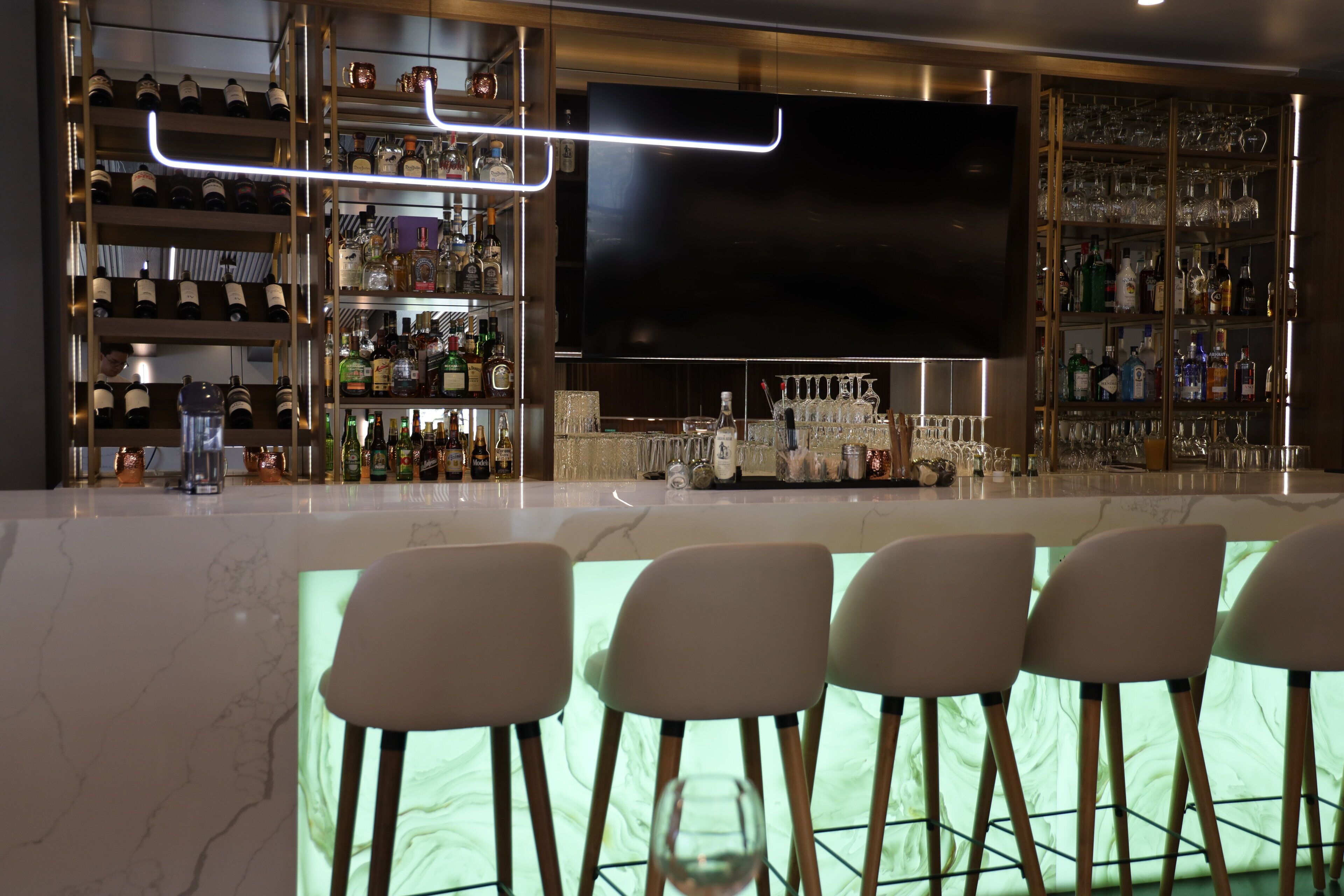 Una zona de bar con una gran pantalla de televisión, estantes con botellas de licor y taburetes.