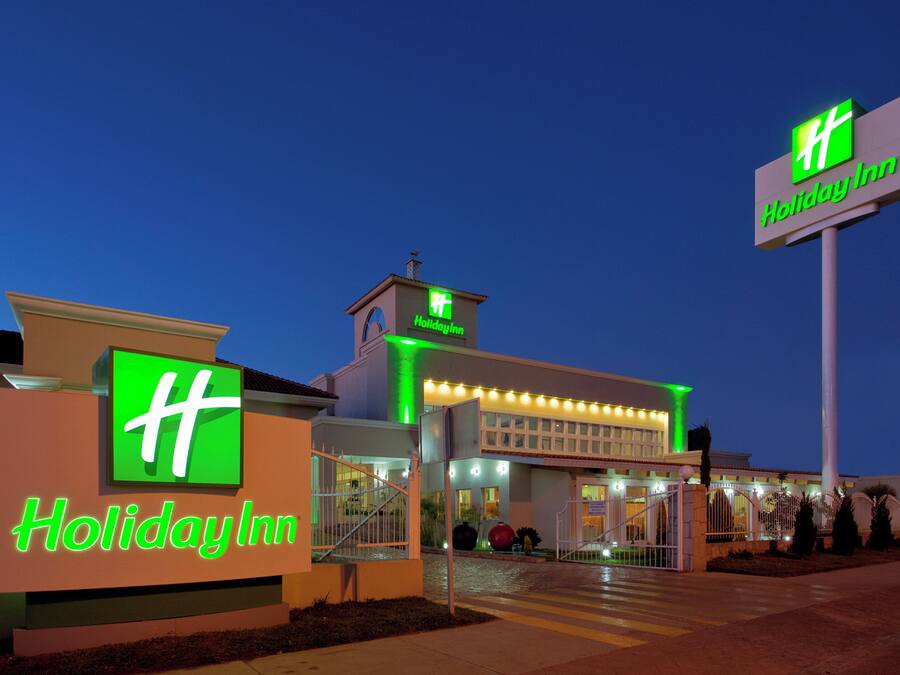 Un hotel Holiday Inn con un logotipo verde destacado, una entrada bien iluminada y un cielo despejado.