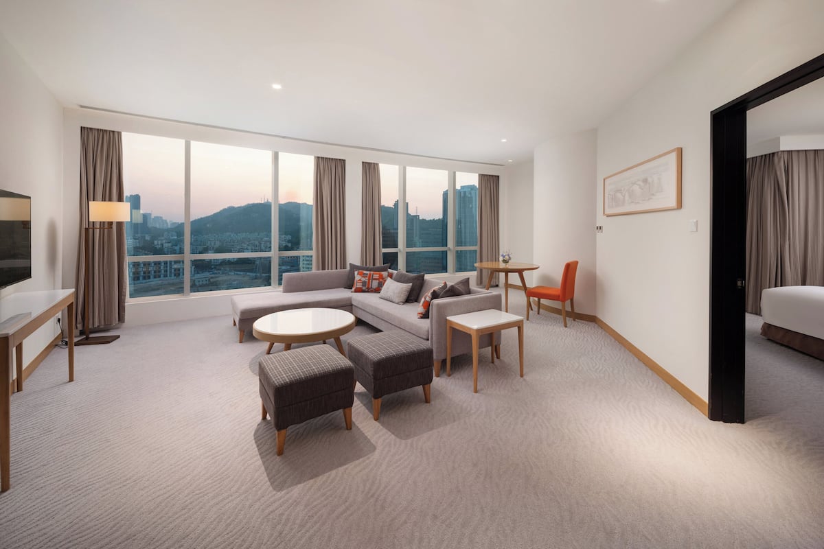 Ein modernes Hotelzimmer mit großem Fenster, einer Couch, einem Esstisch und einem Fernseher.