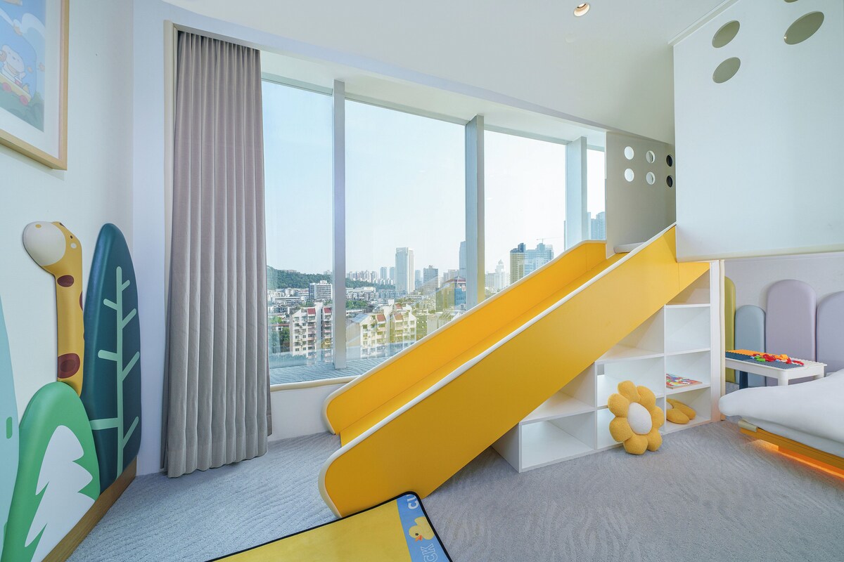 A children's play area with a slide, a large window with a city view, and a bookshelf with toys.