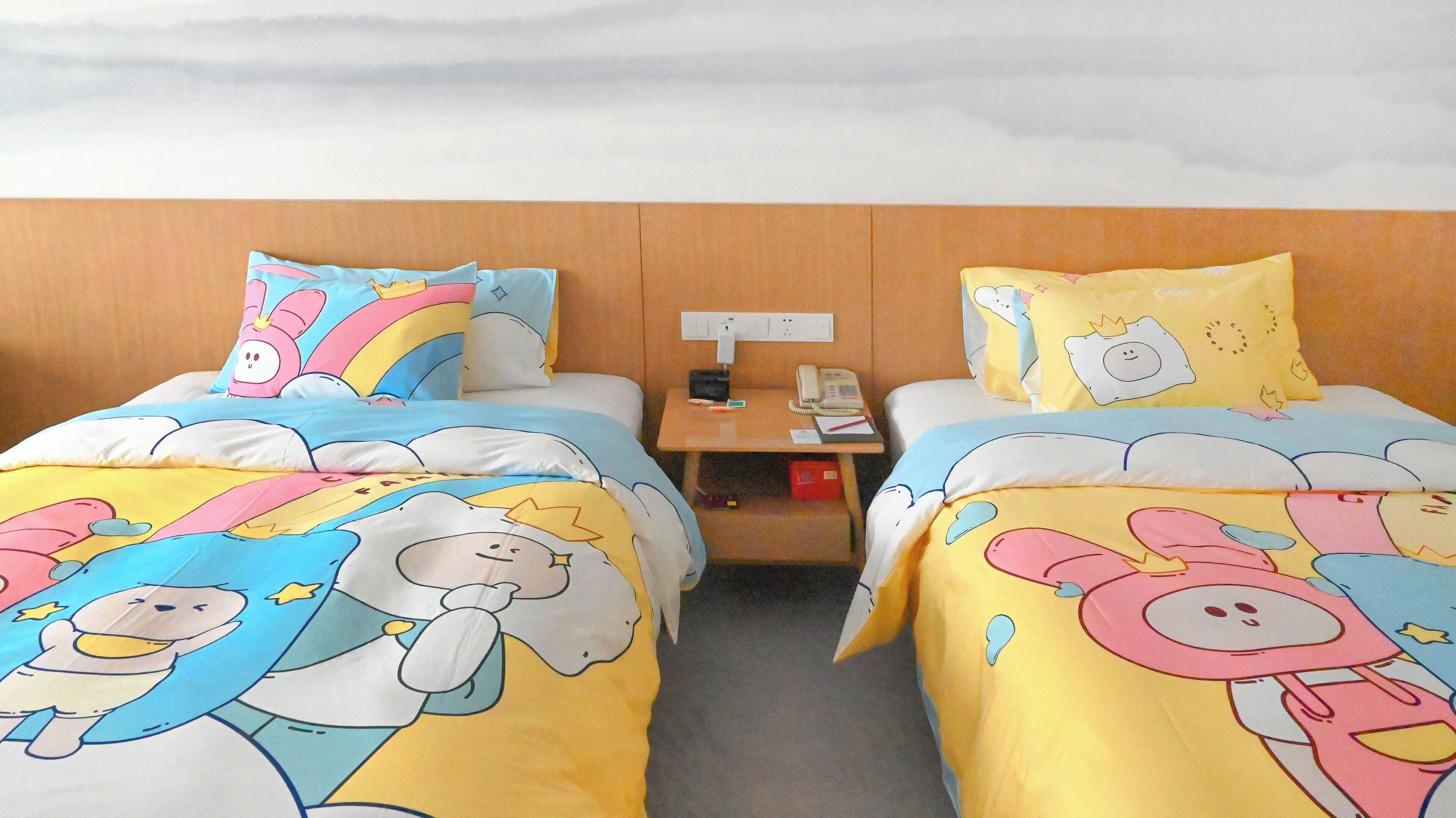 A hotel room with two beds, each with colorful bedding featuring cartoon characters. There is a nightstand with a phone and a lamp between the beds.