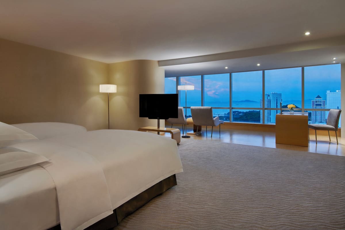Ein Hotelzimmer mit einem großen Bett, einem Fernseher, einem Schreibtisch und einem Sessel. Große Fenster bieten einen Blick auf die Stadt.