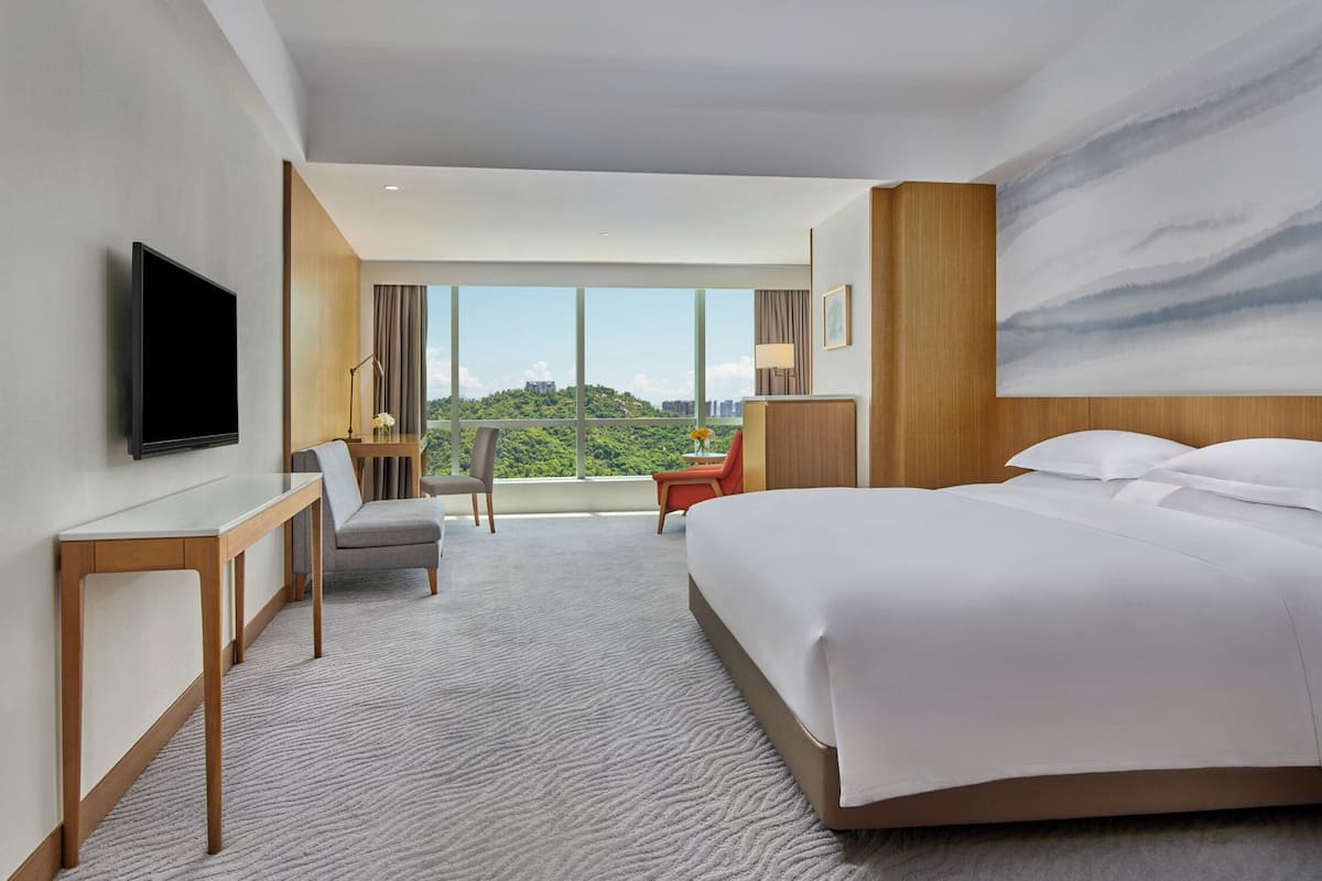 Ein Hotelzimmer mit Bett, Schreibtisch, Stuhl, Fernseher und großem Fenster mit Blick auf die Stadt.