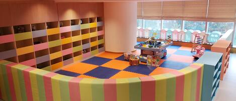 A children's play area with a colorful padded playpen, a play table with toys, and a window with a view of trees.
