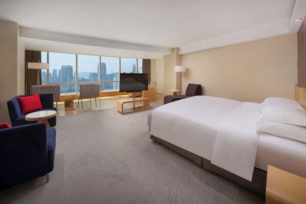 A modern hotel room with a large bed, a seating area with chairs and a table, a flat-screen TV, and a city view.