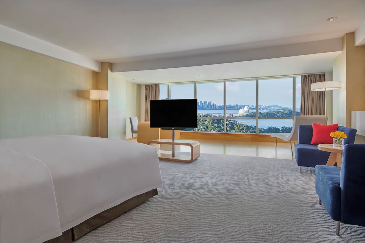 Ein Hotelzimmer mit einem großen Bett, einem Fernseher, einem Schreibtisch und Blick auf eine Stadtlandschaft.