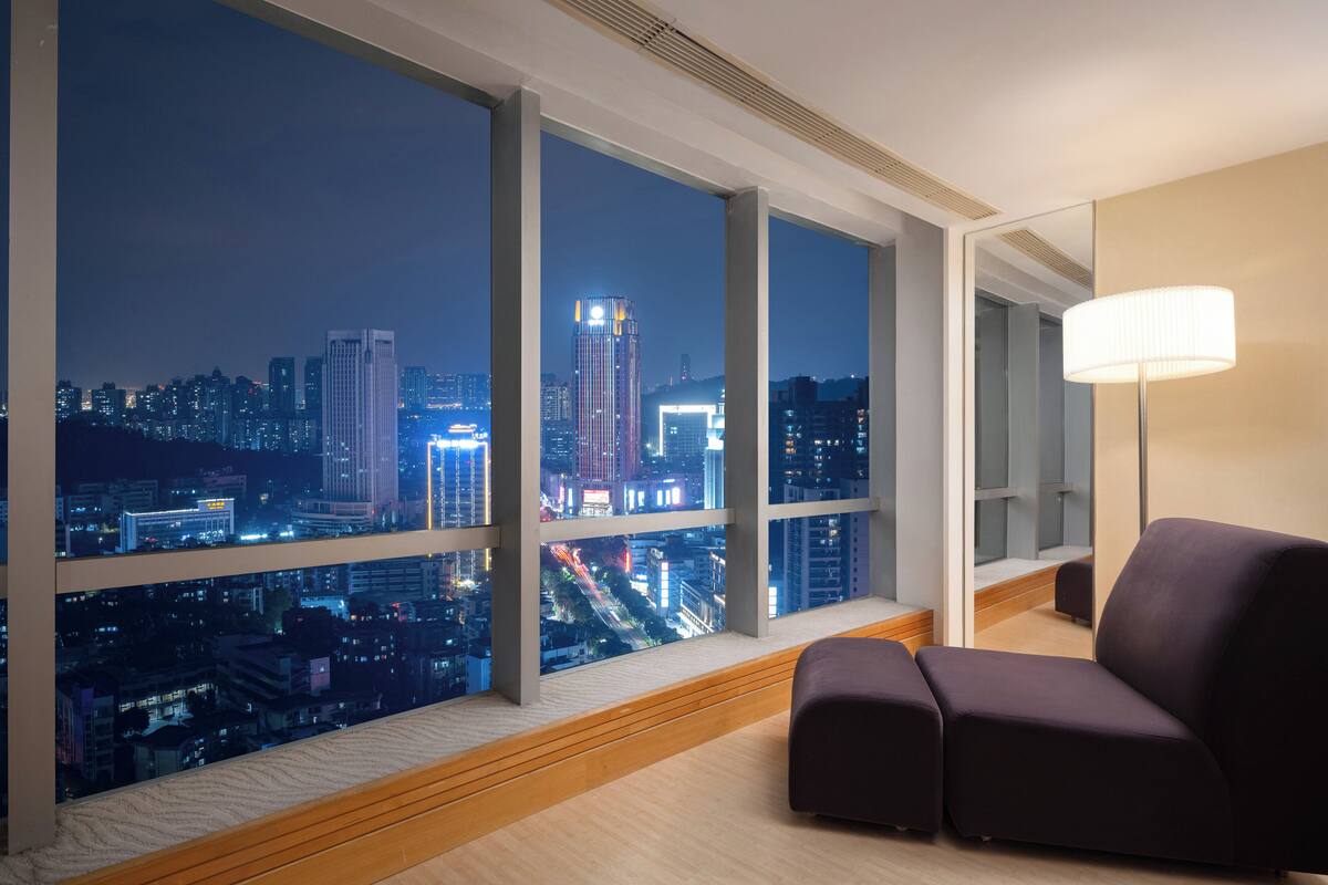A modern hotel room with large windows offering a nighttime city view, a comfortable dark-colored sofa, and a floor lamp.