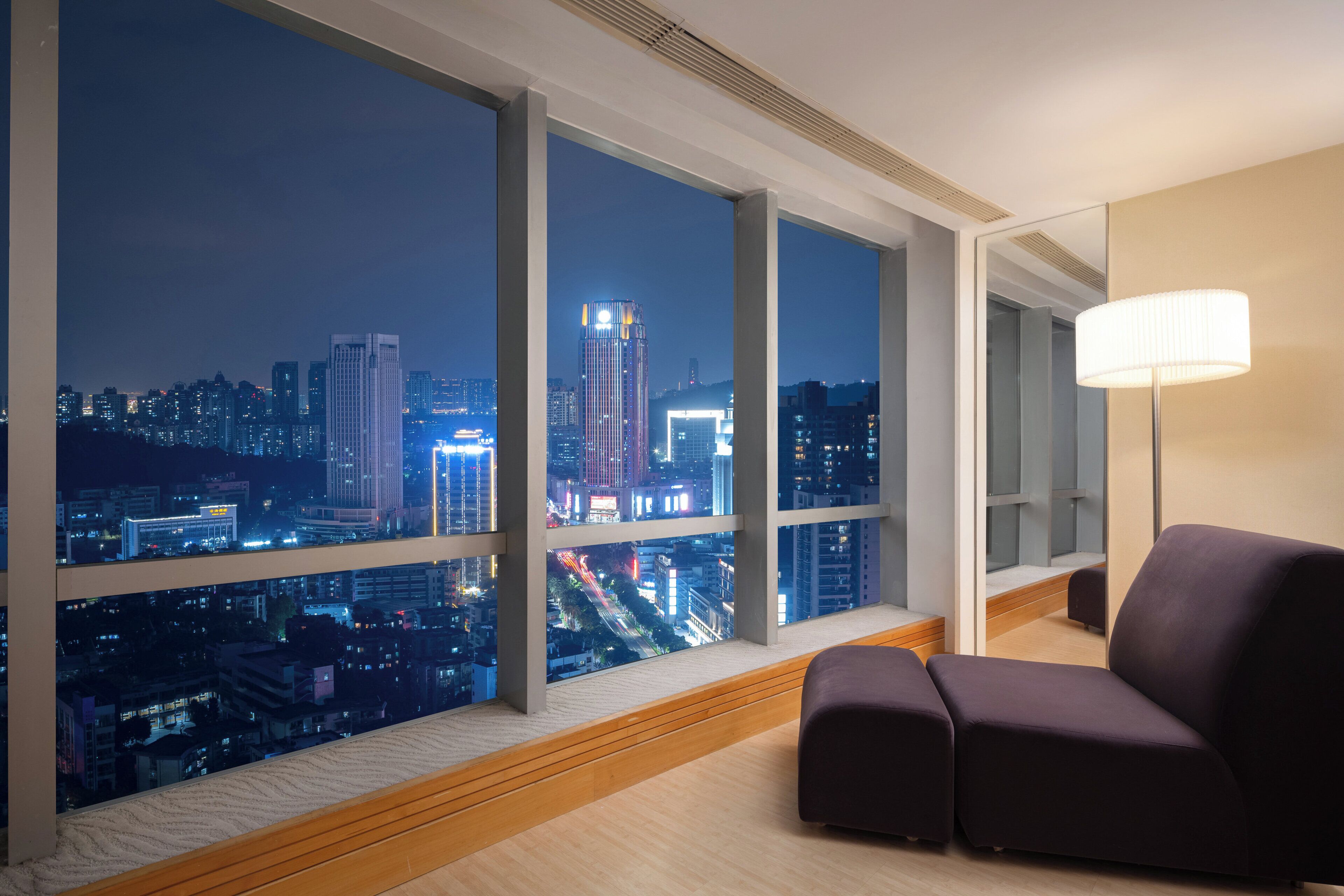 Une chambre d’hôtel moderne dotée de grandes fenêtres offrant une vue sur la ville la nuit, d’un canapé confortable de couleur sombre et d’une lampe sur pied.