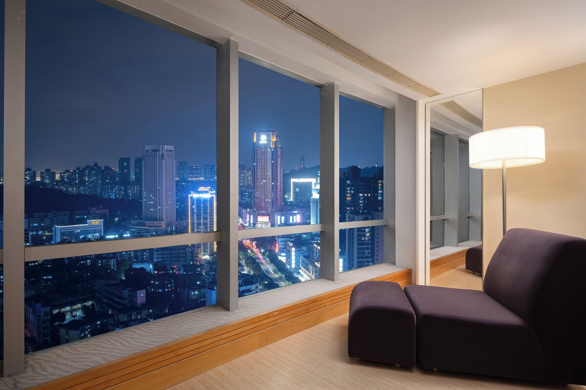 A modern hotel room with large windows offering a nighttime city view, a comfortable dark-colored sofa, and a floor lamp.