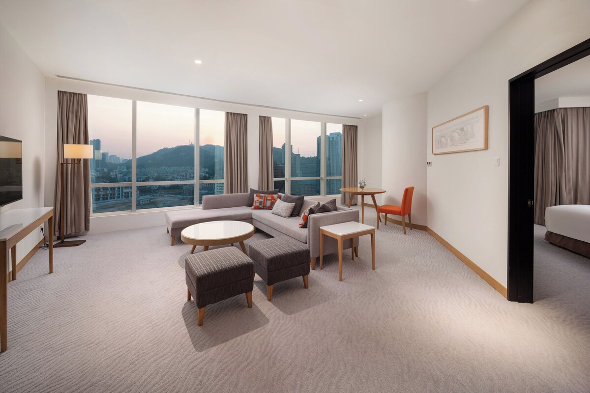 A modern hotel room with a large window, a sofa, a dining table, and a TV.