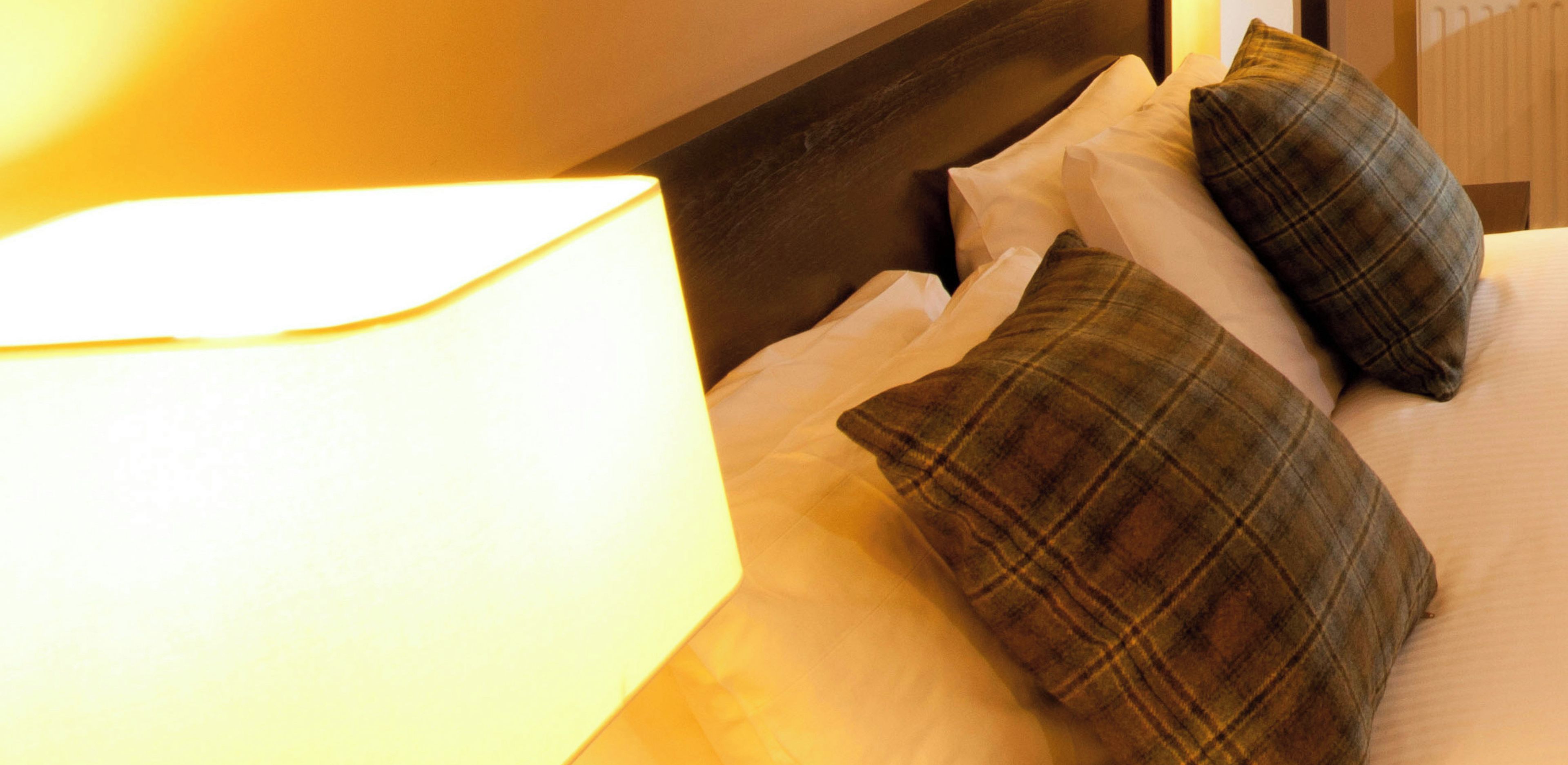 A bed with white linens and two plaid pillows, a bedside lamp, and a wooden headboard.