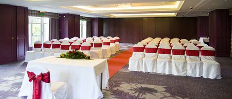 A conference room with rows of white chairs with red sashes, a table with a white tablecloth, and a carpeted floor.