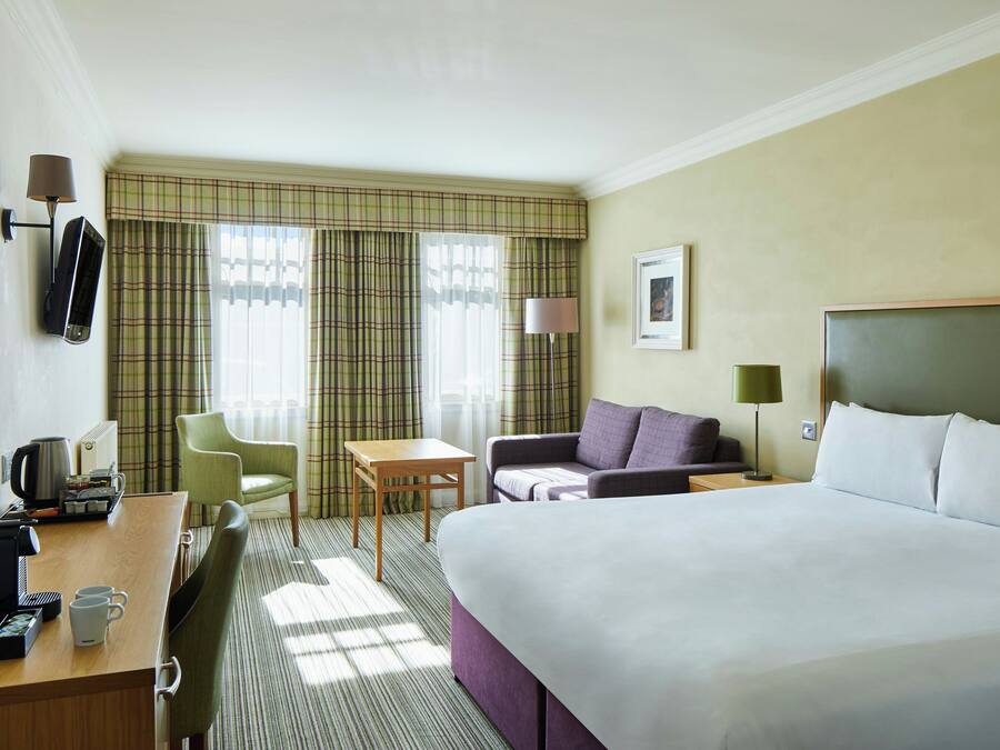 Ein Hotelzimmer mit Bett, Schreibtisch mit Telefon, Stuhl, Tisch und einem Fenster mit Vorhängen.