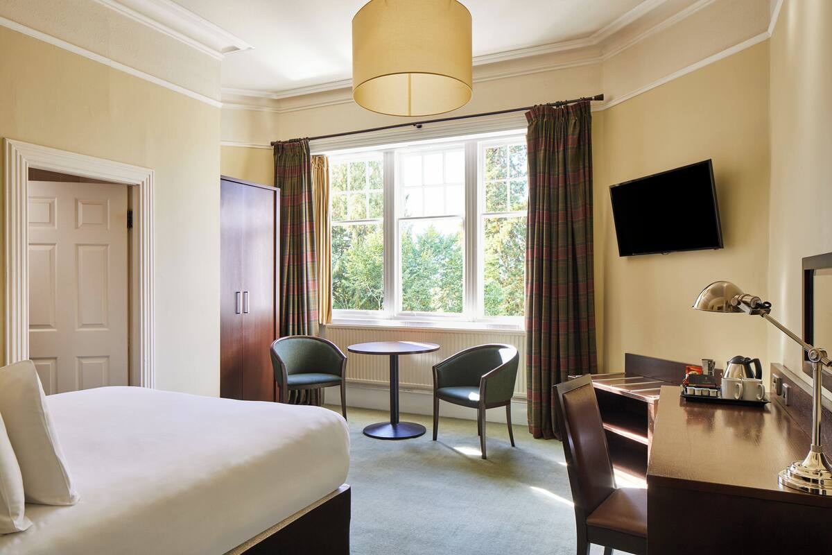 A hotel room with a bed, two chairs, a small round table, a desk with a lamp, a television, and a window with curtains.
