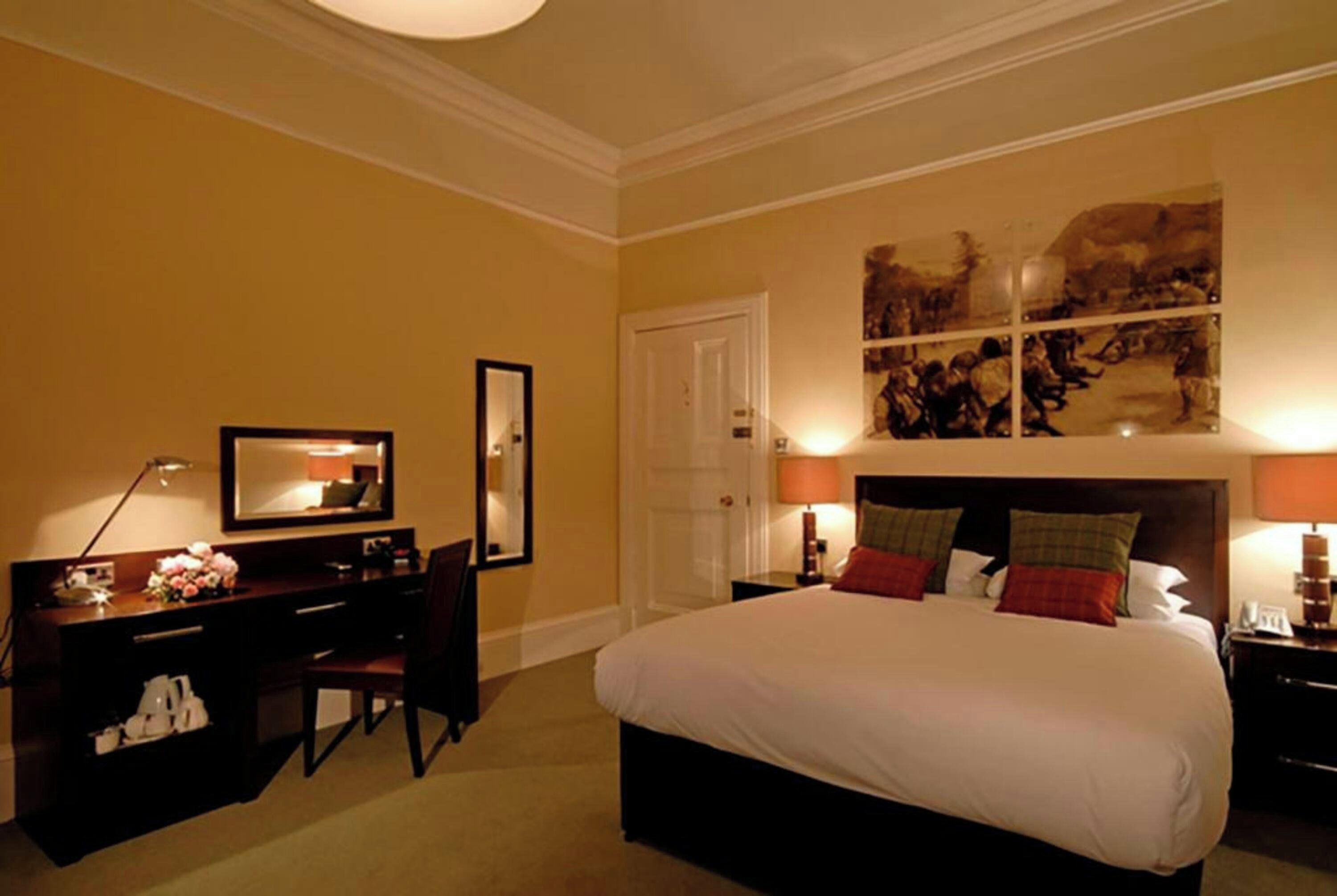 Een hotelkamer met een bed, nachtkastjes, een bureau met stoel, een spiegel en kunst aan de muur.