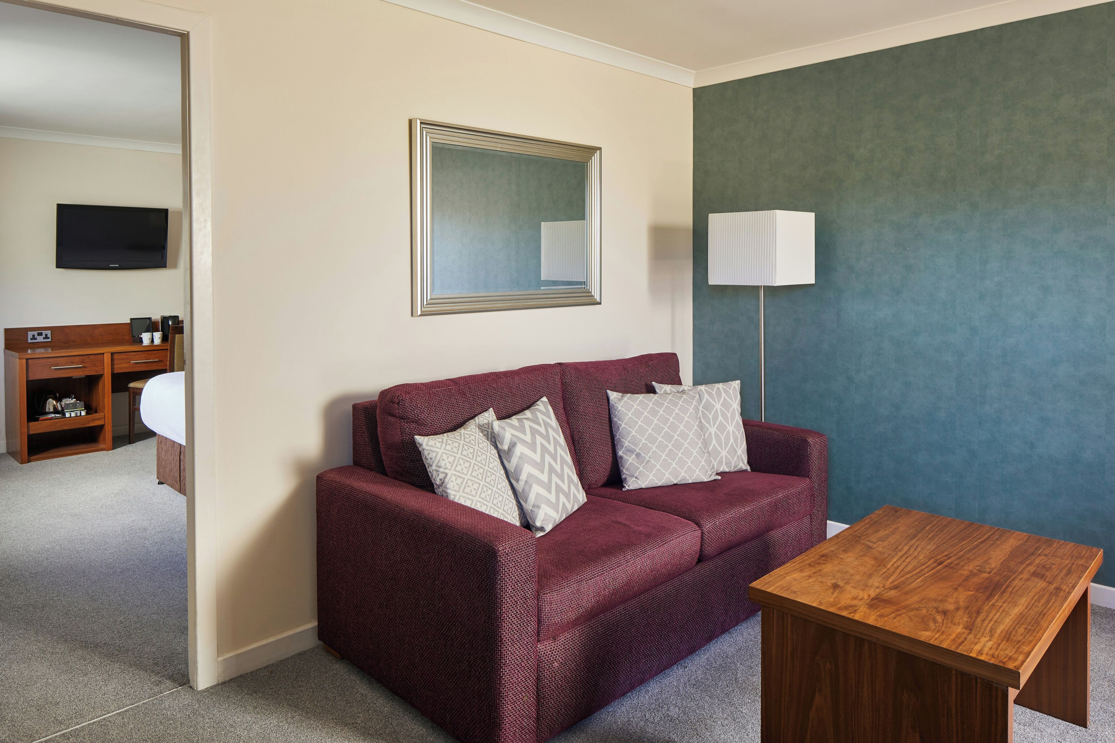 Ein Hotelzimmer mit einem roten Sofa, einem Holztisch, einem Flachbildfernseher, einem Nachttisch mit Lampe und einem Spiegel an der Wand.