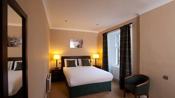 Quarto de hotel com cama, duas mesas de cabeceira com abajures, uma cadeira, uma janela com cortinas e um quadro na parede.