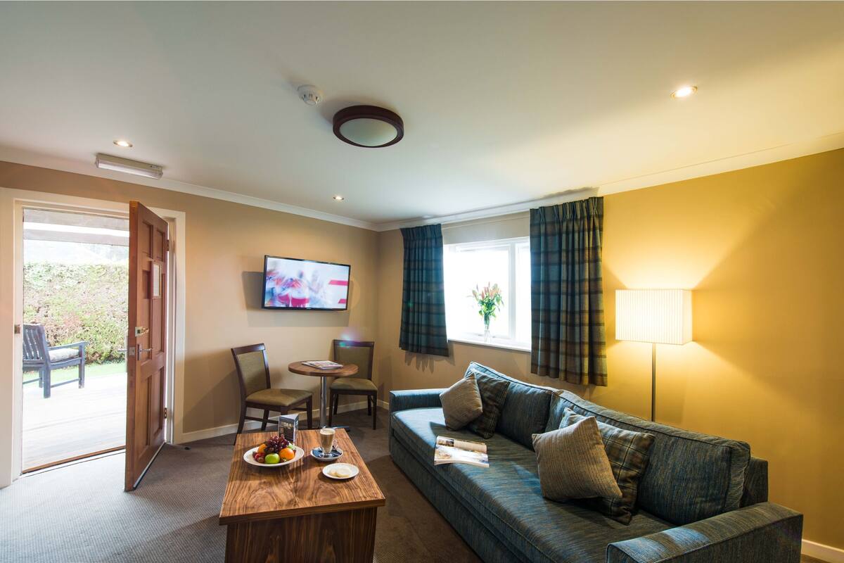 A living room with a sofa, a coffee table with fruits and a drink, a TV on the wall, and a window with curtains.