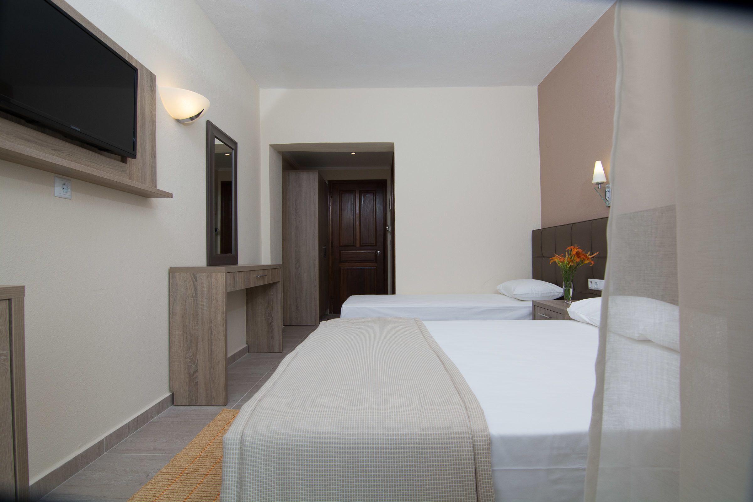 Una habitación de hotel con dos camas, un televisor montado en la pared, un escritorio y un espejo.