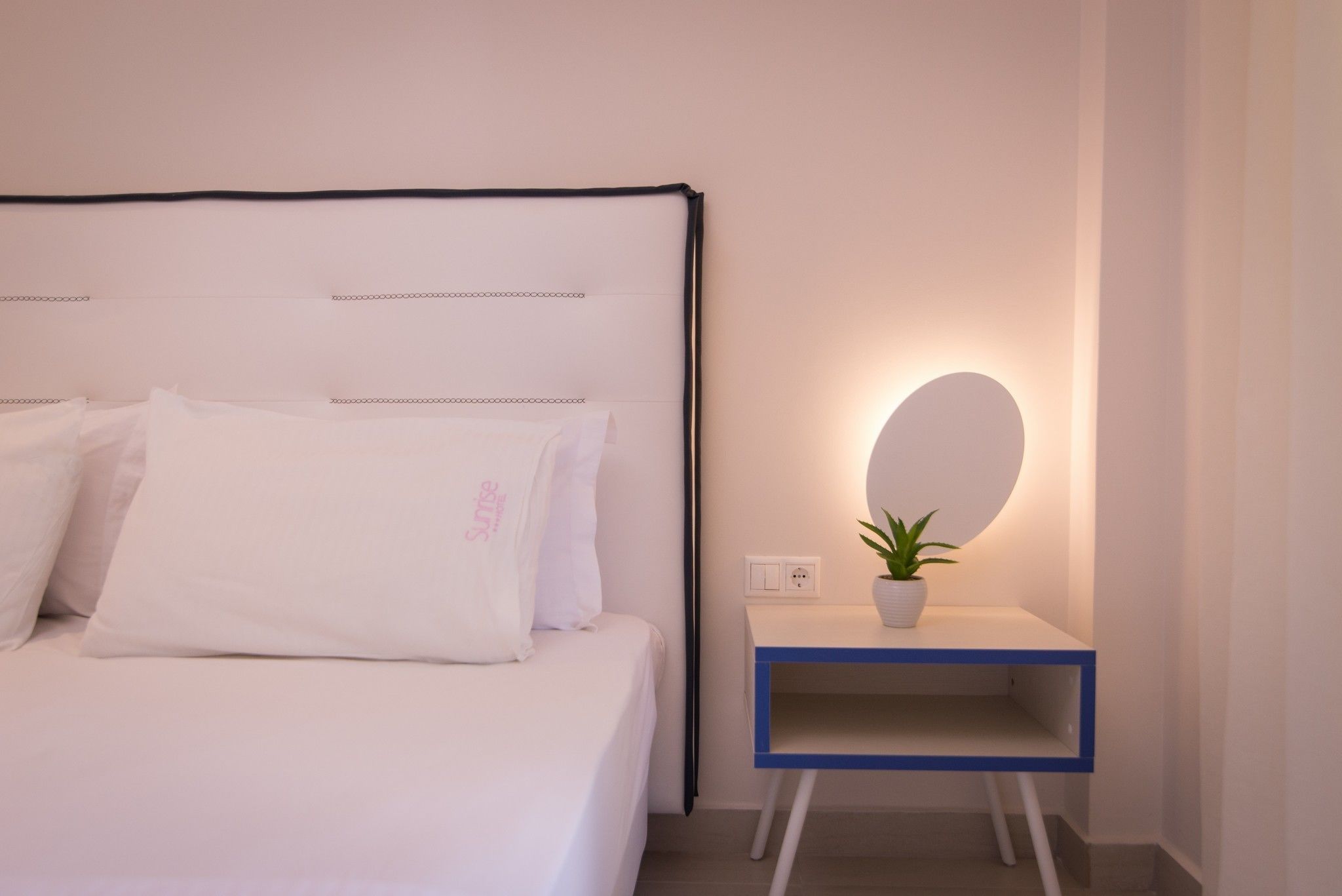 A neatly made bed with white bedding and pillows, a bedside table with a potted plant, and a wall-mounted mirror.