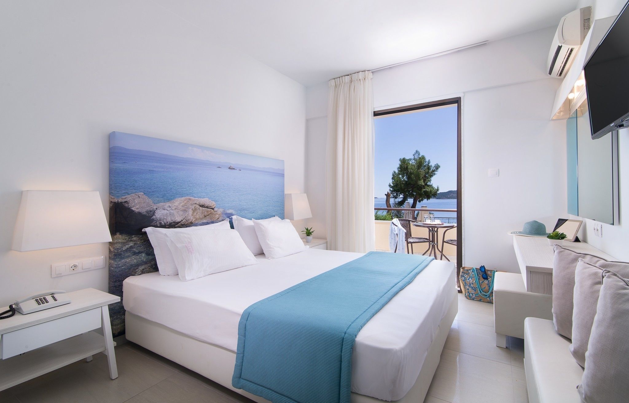 A hotel room with a bed, a desk, a chair, a TV, and a balcony with a view of the sea.