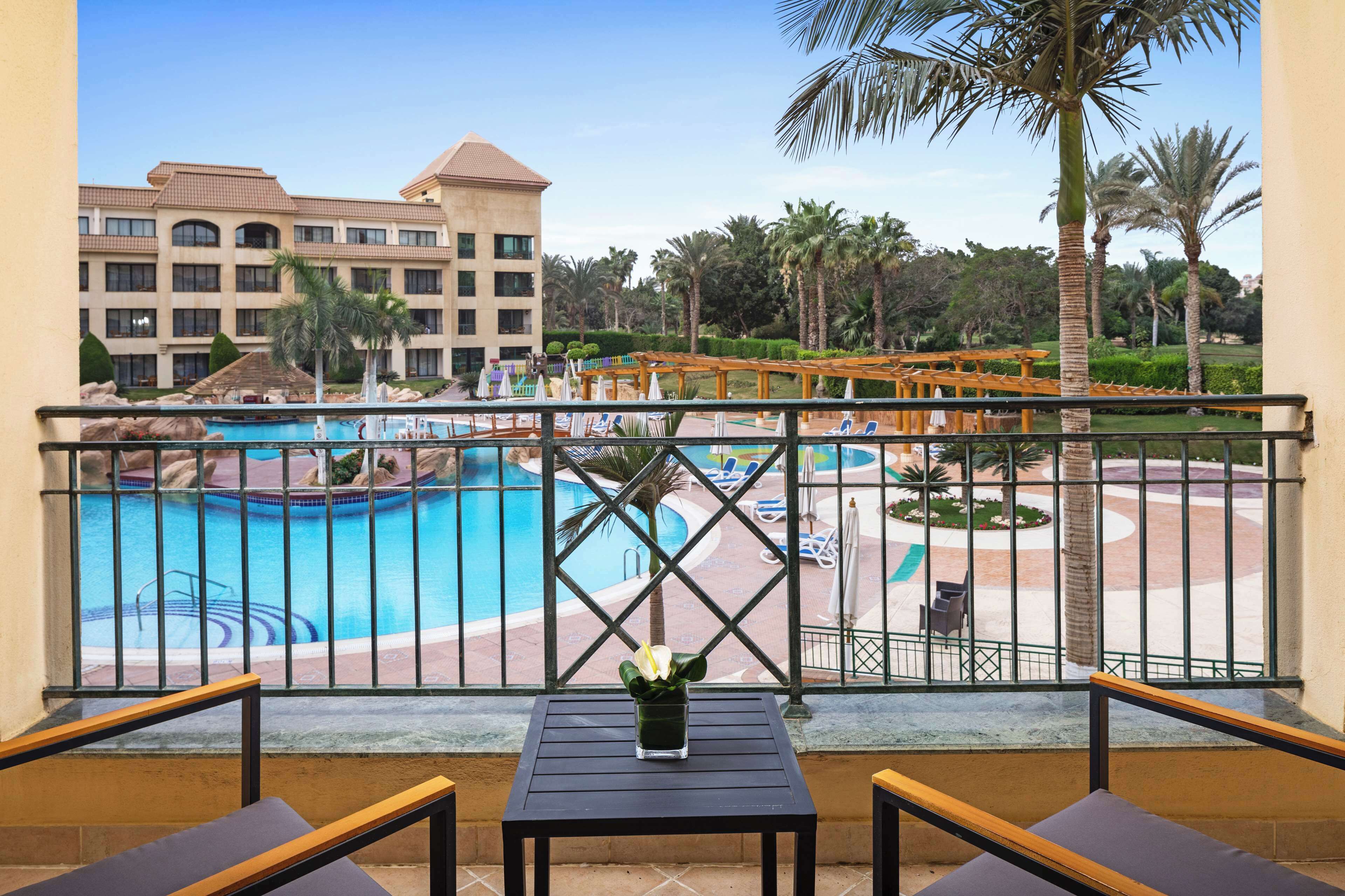 A balcony with a view of a swimming pool, palm trees, and a hotel building.