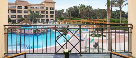 A balcony with a view of a swimming pool, palm trees, and a hotel building.