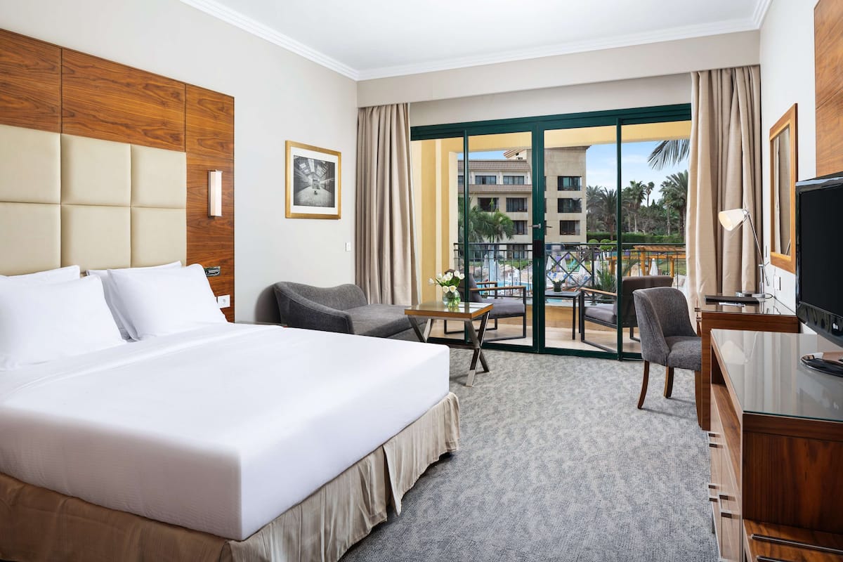 A hotel room with a large bed, a desk, a chair, and a balcony with a view of buildings and palm trees.