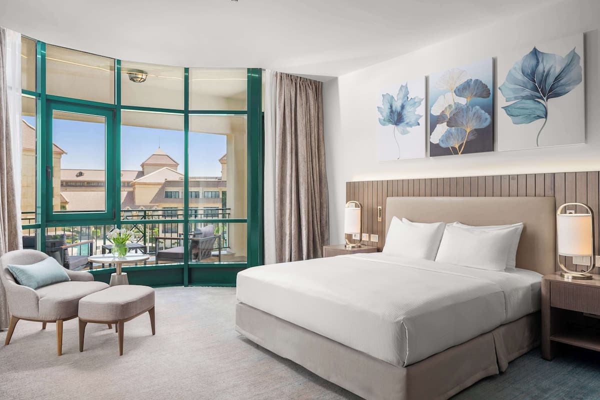 A hotel room with a large bed, a chair, a small table, and a view of buildings through a large window.