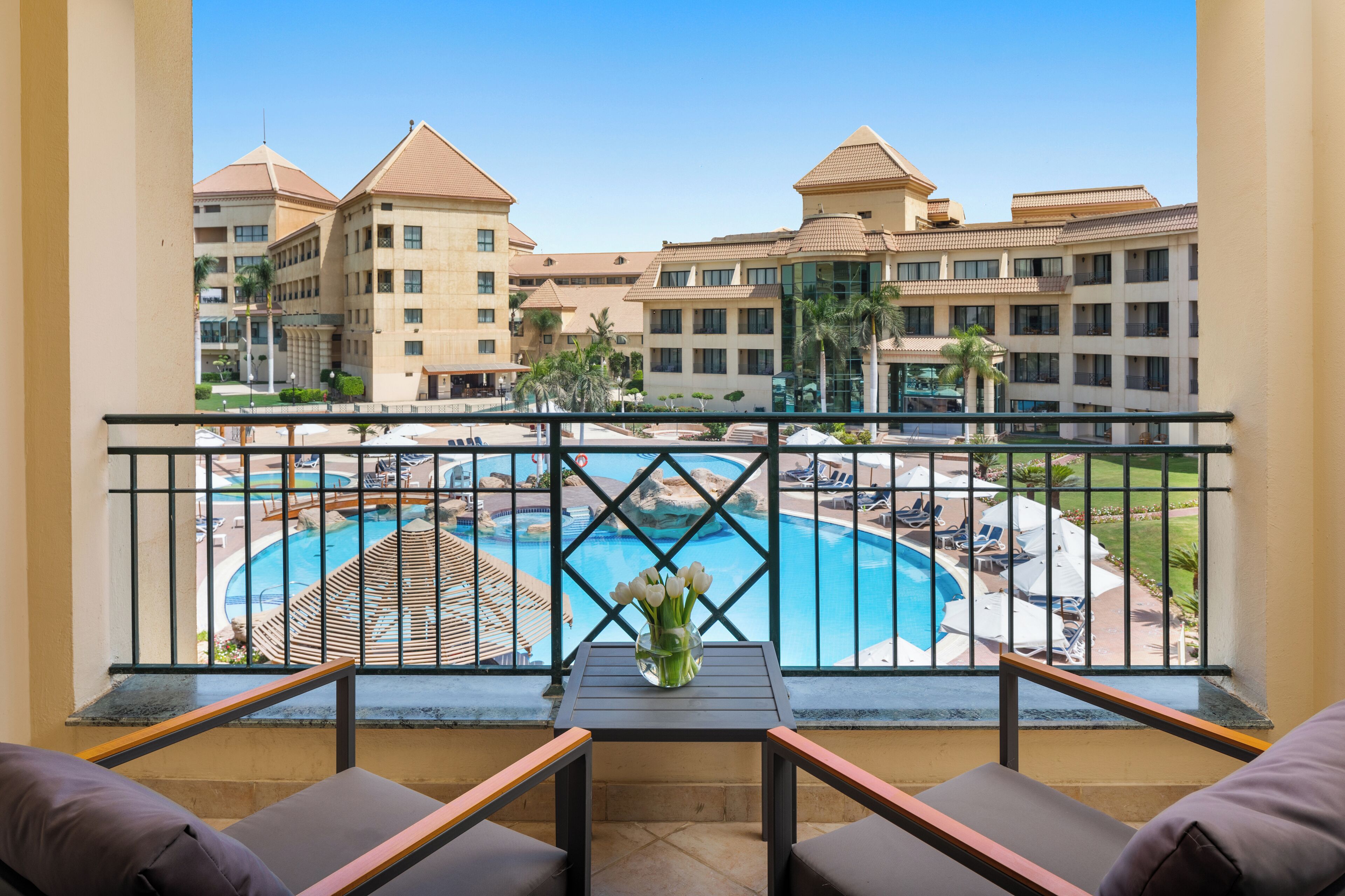 A balcony with a view of a swimming pool, lounge chairs, and a table with a vase of flowers.