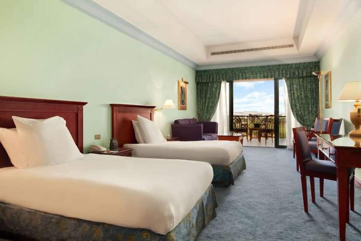 A hotel room with two beds, a desk, and a chair. There is a view of a balcony with a table and chairs.