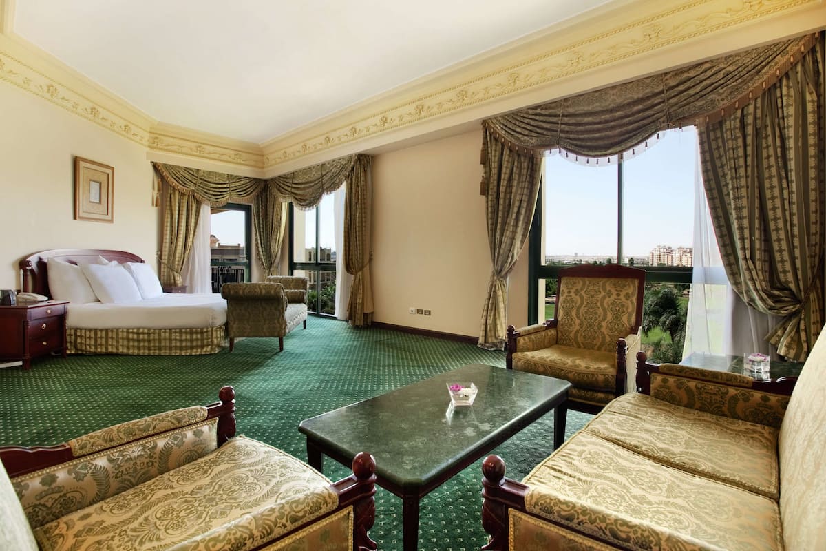 A hotel room with a green carpet, a bed, a sofa, a chair, a table, and large windows with curtains.