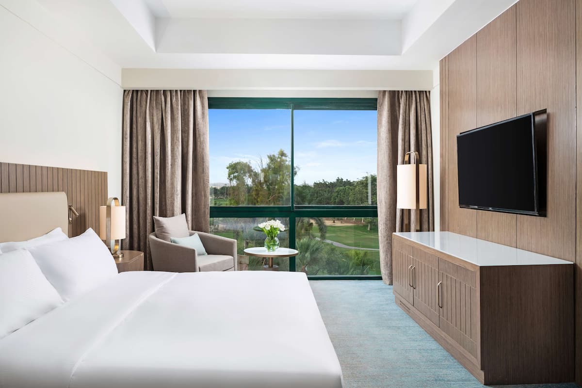 A hotel room with a large bed, a TV mounted on the wall, a small table with a lamp, a chair, and a view of a golf course through the window.